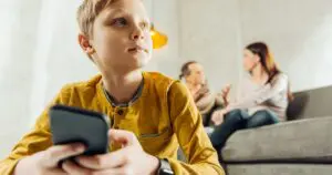 A child uses their smartphone while their parents talk on the sofa in the background.