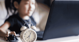 A child sits using a laptop in the background, blurred behind an alarm clock in the foreground.