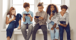 5 children sitting on a windowsill looking at their individual smartphones.