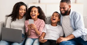 A family of four - woman, girl, boy and man - laughing while sitting in the sofa with their devices