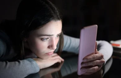 A girl in a dark room stares at her smartphone.