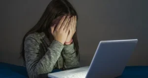 A girl sits in front of a laptop in the dark with her face covered by her hands.