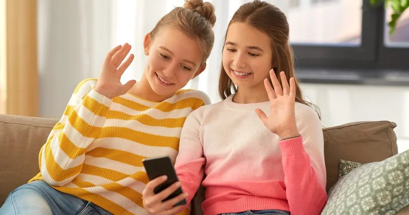 Two teens wave at someone on their smartphone.