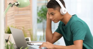 A teen boy wears headphones while browsing on a laptop.