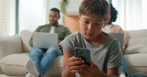 A boy sits on the floor using his smartphone near his parents.